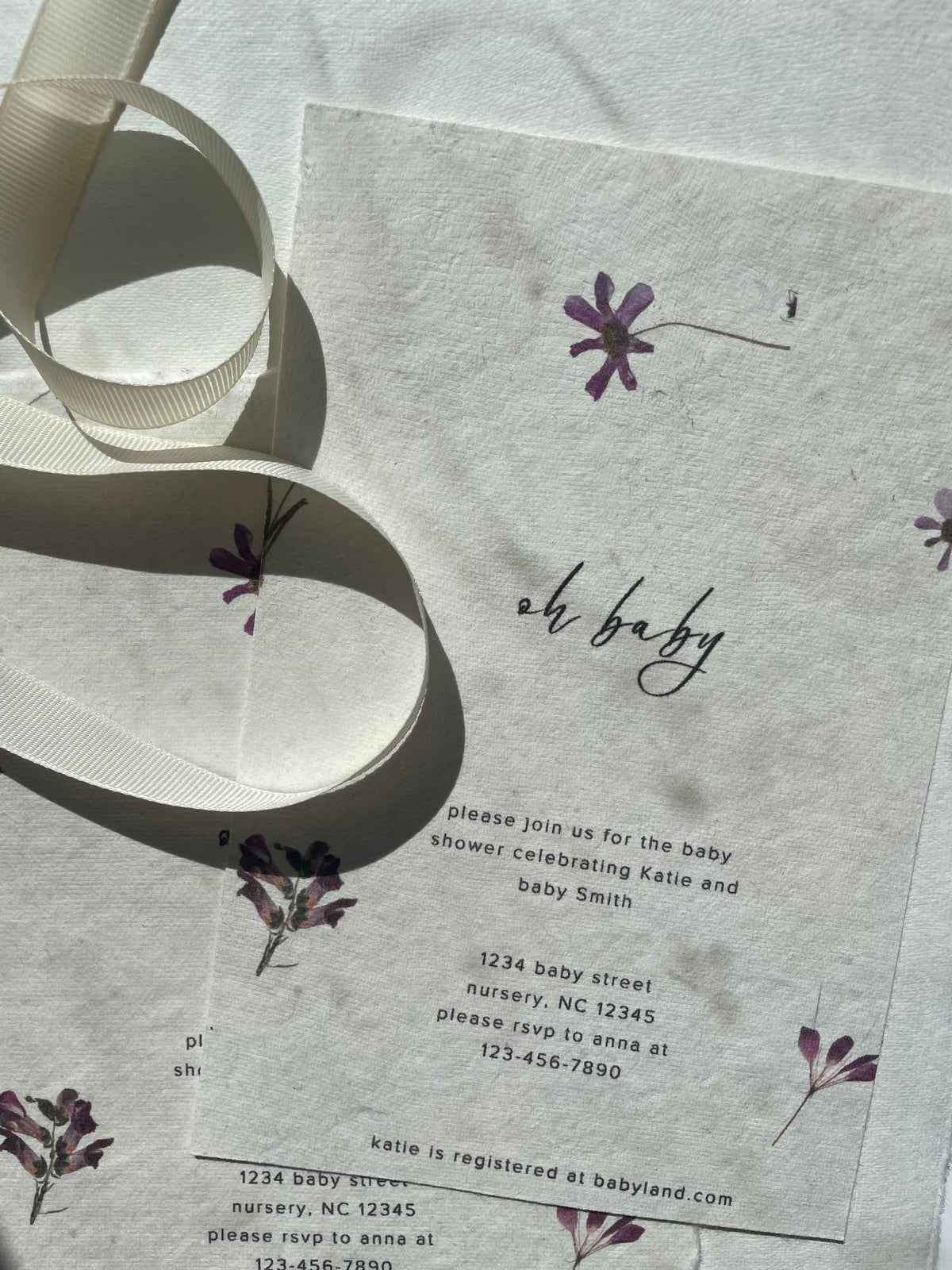 A close-up of a baby shower invitation card with a white ribbon draped across it. The card has a textured handmade cotton off-white background with delicate purple floral illustrations and black text, including the phrase 'oh baby'.