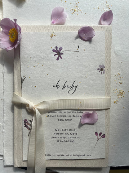 A flat-lay of a baby shower invitation card with a cream ribbon tied in a bow, decorated with dried lavender flowers and scattered petals on a textured white background.