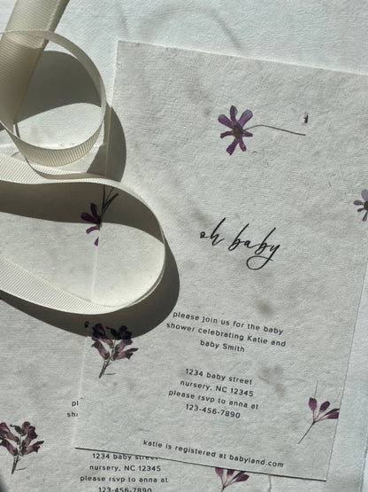 A close-up of a baby shower invitation card with a white ribbon draped across it. The card has a textured handmade cotton off-white background with delicate purple floral illustrations and black text, including the phrase 'oh baby'.